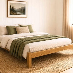 May include: A wooden bed frame with a neutral-colored comforter and pillows. A green throw blanket is draped across the foot of the bed. The bed sits on a woven jute rug. A framed landscape painting hangs on the wall.