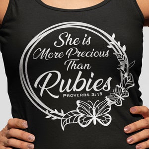 Puede incluir: Camiseta de tirantes negra con un diseño gráfico blanco que dice "She is More Precious Than Rubies" con una corona de flores y un diseño de mariposa. El texto "Proverbs 3:15" también está incluido.