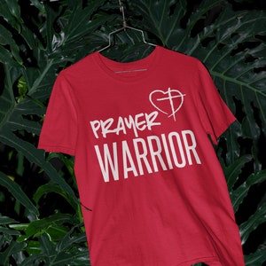 K&ouml;nnte beinhalten: Rotes T-Shirt mit wei&szlig;em Text "PRAYER WARRIOR" und einem wei&szlig;en Herz mit einem Kreuz darin.
