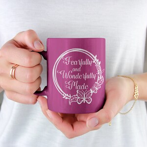 May include: A purple ceramic mug with a white design that says "Fearfully and Wonderfully Made" in a decorative font. The design is surrounded by a wreath of flowers and butterflies.