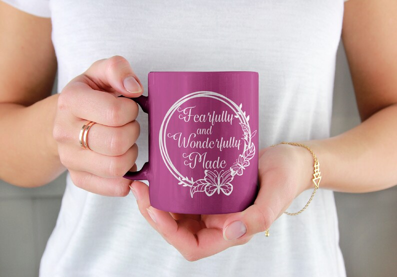 May include: A purple ceramic mug with a white design that says "Fearfully and Wonderfully Made" in a decorative font. The design is surrounded by a wreath of flowers and butterflies.