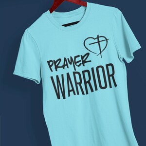 K&ouml;nnte beinhalten: Ein hellblaues T-Shirt mit dem Text "PRAYER WARRIOR" und einem Herz mit einem Kreuz darin.