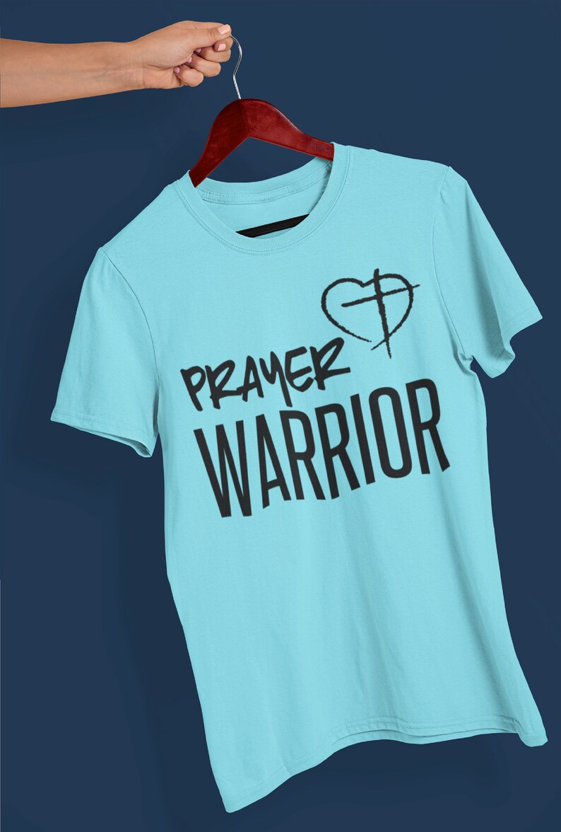 K&ouml;nnte beinhalten: Ein hellblaues T-Shirt mit dem Text "PRAYER WARRIOR" und einem Herz mit einem Kreuz darin.