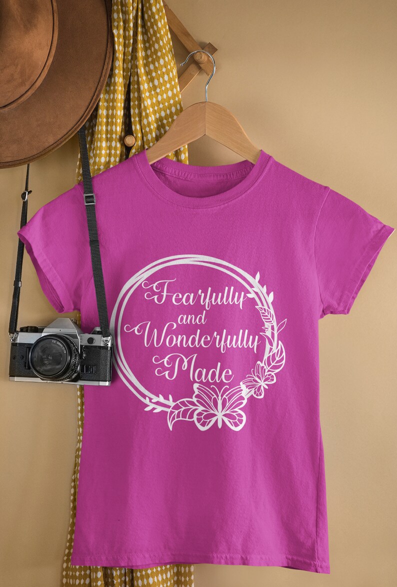 May include: A pink t-shirt with a white circle design featuring the text "Fearfully and Wonderfully Made" with a floral wreath and butterfly accents.