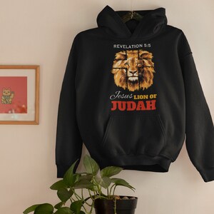 Może przedstawiać: Czarna bluza z kapturem z grafiką przedstawiającą twarz lwa z nadrukiem "Revelation 5:5 Jesus Lion of Judah" na przodzie.