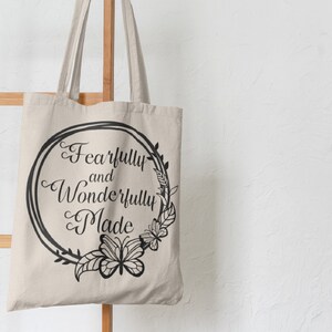 May include: A beige canvas tote bag with black handles and a black circular design featuring the text "Fearfully and Wonderfully Made" surrounded by a floral wreath.