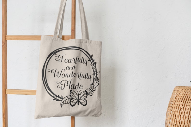 May include: A beige canvas tote bag with black handles and a black circular design featuring the text "Fearfully and Wonderfully Made" surrounded by a floral wreath.