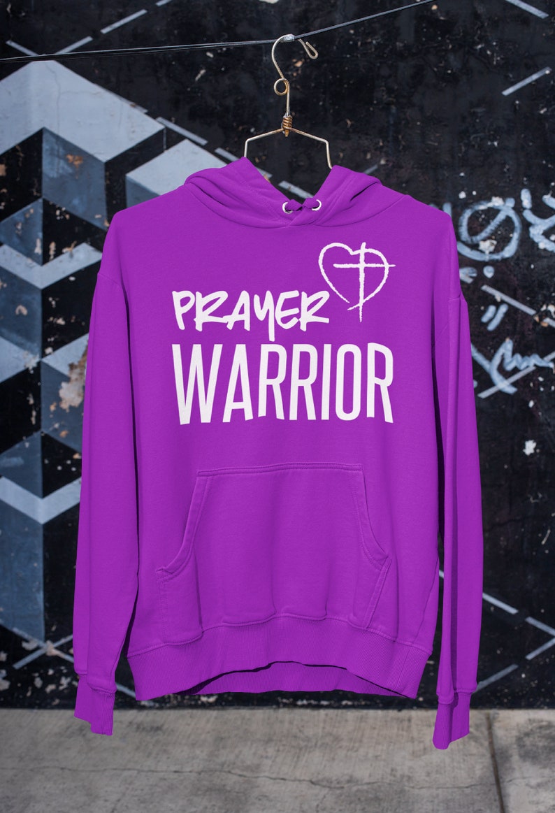 K&ouml;nnte beinhalten: Lilafarbener Hoodie mit einem wei&szlig;en Aufdruck, der "PRAYER WARRIOR" und ein Herz mit einem Kreuz darin zeigt.