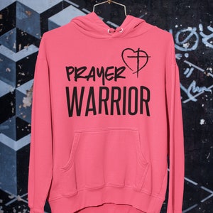 K&ouml;nnte beinhalten: Ein rosa Hoodie mit einem schwarzen Aufdruck, der "PRAYER WARRIOR" mit einem Herz und einem Kreuz zeigt.