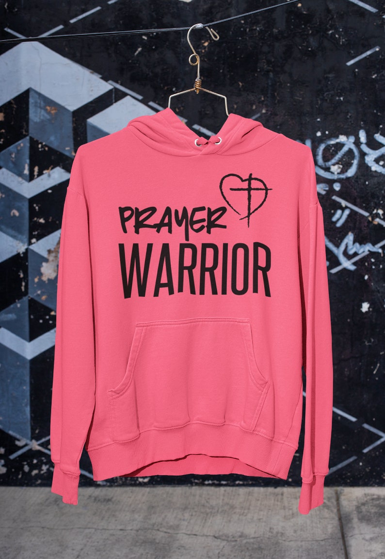 K&ouml;nnte beinhalten: Ein rosa Hoodie mit einem schwarzen Aufdruck, der "PRAYER WARRIOR" mit einem Herz und einem Kreuz zeigt.