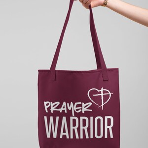 K&ouml;nnte beinhalten: Eine weinrote Tragetasche mit wei&szlig;em Text, der "PRAYER WARRIOR" lautet, und einem wei&szlig;en Herzen mit einem Kreuz darin.