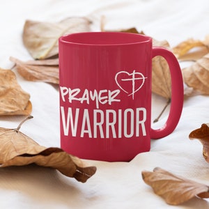 K&ouml;nnte beinhalten: Eine rote Keramiktasse mit einem wei&szlig;en Design, auf dem "Prayer Warrior" mit einem Herz- und Kreuzsymbol steht.