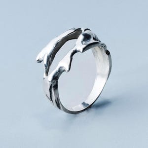 Peut inclure: Une bague en argent au design organique et fluide. La bague présente un aspect texturé, semblable à de la fonte, avec des formes et des courbes irrégulières. Le métal a une finition polie, reflétant la lumière. Le design est abstrait et moderne.