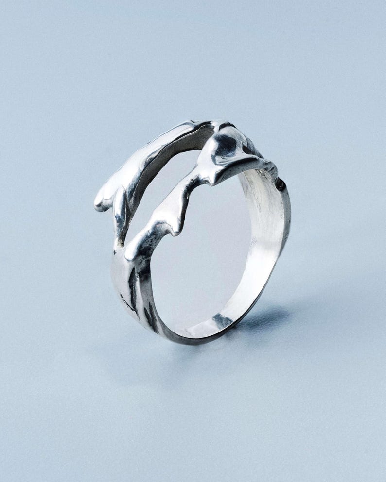 May include: A silver ring with an organic, flowing design. The ring features a textured, molten-like appearance, with irregular shapes and curves. The metal has a polished finish, reflecting light. The ring's design is abstract and modern, suitable for everyday wear.