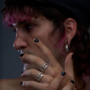 May include: A close-up of a person's hands adorned with silver rings and a chain bracelet. The rings feature a unique, spiky design. The person has black nail polish and a nose ring. The background is a dark gray.