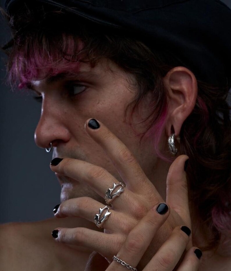 May include: A close-up of a person's hands adorned with silver rings and a chain bracelet. The rings feature a unique, spiky design. The person has black nail polish and a nose ring. The background is a dark gray.