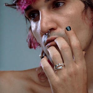 May include: A silver-colored ring with an organic, branch-like design. The ring features an open, irregular shape, with multiple prongs and curves. The ring is worn on a finger.