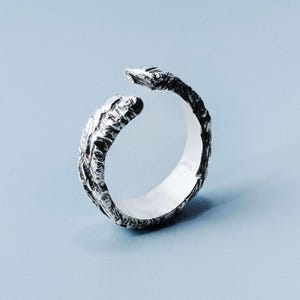 Puede incluir: Un anillo plateado con un diseño orgánico y texturizado. El anillo tiene un diseño abierto, con los extremos de la banda curvándose alejándose uno del otro. La superficie del anillo tiene una textura rugosa, similar a la corteza. El fondo es azul claro liso.