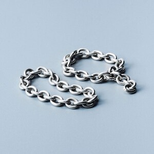 Può includere: Due braccialetti a catena argentati sono esposti su una superficie azzurra. I braccialetti sono composti da piccole maglie ovali. Un braccialetto è di forma circolare, mentre l'altro è parzialmente slacciato, creando un design lineare.