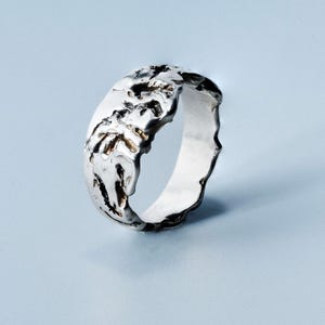 Può includere: Un anello d'argento con un design strutturato e organico. L'anello ha una superficie irregolare con un aspetto ruvido, quasi fuso. L'interno dell'anello è liscio e lucido, in contrasto con la texture ruvida dell'esterno. L'anello è posto su uno sfondo azzurro.