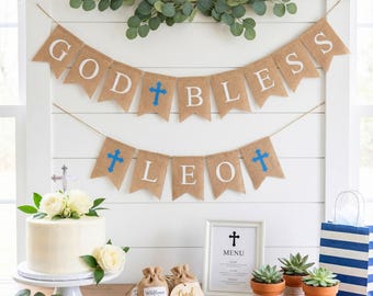 May include: A celebratory scene with a banner reading "GOD BLESS LEO" and a cake. The banner is made of burlap with white letters and blue crosses. A white cake with flowers sits on a white cake stand. Other decorations include succulents and a menu.