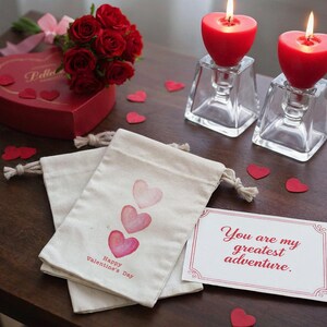 May include: A romantic Valentine's Day scene with red heart-shaped candles in glass holders, a bouquet of red roses, and a heart-shaped box. Two drawstring bags with watercolor hearts and the text "Happy Valentine's Day" are also present, along with a card that reads "You are my greatest adventure."