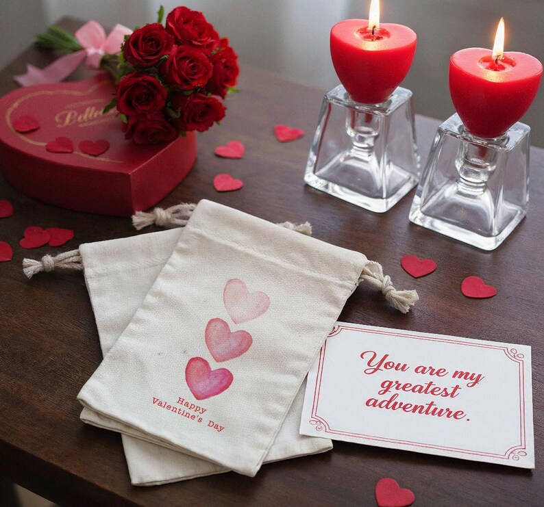 May include: A romantic Valentine's Day scene with red heart-shaped candles in glass holders, a bouquet of red roses, and a heart-shaped box. Two drawstring bags with watercolor hearts and the text "Happy Valentine's Day" are also present, along with a card that reads "You are my greatest adventure."