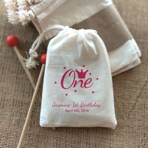 May include: White cotton drawstring bag with a pink crown and the word "One" printed on it. The bag also has the text "Jasmine 1st Birthday April 4th, 2016" printed on it.