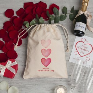 May include: A Valentine's Day gift set featuring a drawstring bag with three pink hearts and the text "Happy Valentine's Day." The set includes a red heart-shaped box, champagne, rose petals, and a greeting card.