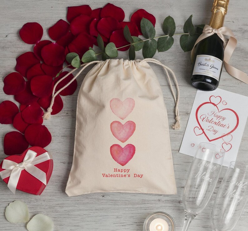 May include: A Valentine's Day gift set featuring a drawstring bag with three pink hearts and the text "Happy Valentine's Day." The set includes a red heart-shaped box, champagne, rose petals, and a greeting card.