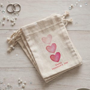 May include: A stack of beige fabric gift bags with three watercolor hearts in shades of pink. The words "Happy Valentine's Day" are printed in red. The bags have drawstring closures.