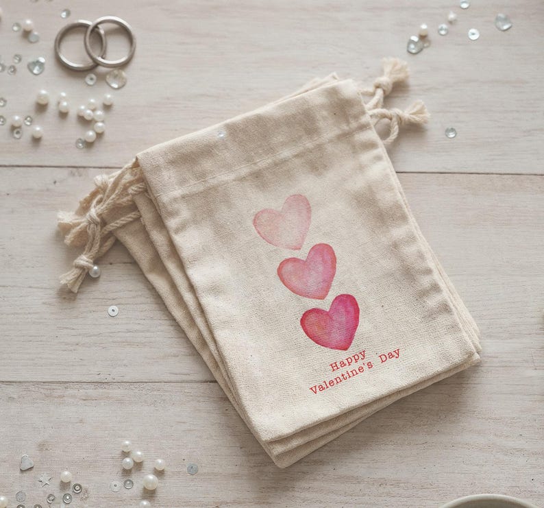 May include: A stack of beige fabric gift bags with three watercolor hearts in shades of pink. The words "Happy Valentine's Day" are printed in red. The bags have drawstring closures.