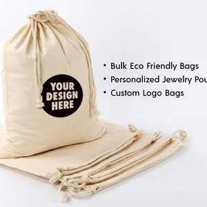 May include: A stack of six natural cotton drawstring bags with a white background. The top bag has a black circle with the text "YOUR DESIGN HERE". The text below the image reads: "Bulk Eco Friendly Bags", "Personalized Jewelry Pouches", and "Custom Logo Bags".