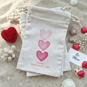 May include: Two off-white drawstring bags with watercolor hearts and the text "Happy Valentine's Day." A red heart-shaped box, seashells, and a card that says "Be Mine" are also present. The items are arranged on sand.
