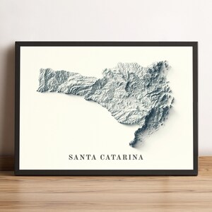 May include: A framed, grayscale 3D topographic map of Santa Catarina, Brazil. The map is set against a cream-colored background, with the state's name printed in a serif font below. The frame is black, and the piece rests on a wooden surface.