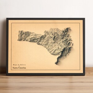 May include: Framed relief map of Santa Catarina, Brazil, in shades of beige and brown. The map is encased in a black frame, with the text "Mapa de Relevo de Santa Catarina" at the bottom left. The map's 3D effect highlights the region's topography.