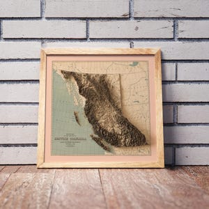 Vintage British Columbia Canada shaded relief map in a wooden frame. Realistic terrain art print for home decor, displayed against a brick wall