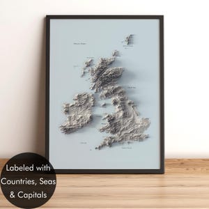 May include: A framed wall art print featuring a three-dimensional relief map of the British Isles in shades of gray against a light blue background. The map displays detailed topography of the landmasses, labeled with names of countries, seas, and capitals.