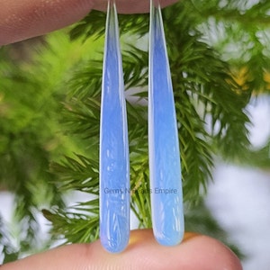 May include: A pair of teardrop-shaped opal earrings with a light blue color. The earrings are hanging from a person's hand, with a green background.