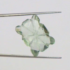 Puede incluir: Una piedra preciosa facetada de color verde claro con forma de estrella de ocho puntas. La piedra preciosa está sujeta por unas pinzas.