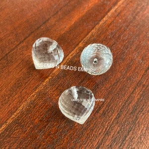May include: Three clear quartz crystal beads, two heart-shaped and one round, with faceted surfaces. The beads are on a wooden surface. "Gems N Beads Empire" is written on the surface.