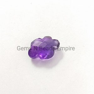 May include: A flower-shaped amethyst gemstone with multiple facets. The gem is a deep purple colour, catching the light. The gemstone is set against a white background. The text "Gems N Beads Empire" is visible.