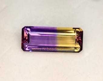 May include: A bi-color gemstone with a purple and yellow gradient. The gemstone is cut into a rectangular shape with facets. The gemstone is approximately 1 inch long.