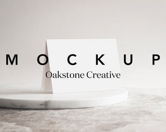 May include: White card mockup on a white marble surface. The card is folded in half and has the text "MOCKUP Oakstone Creative" printed on the front.