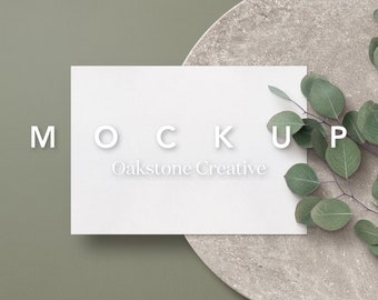 May include: A white card with the text "MOCKUP Oakstone Creative" on a green background with a round, light brown surface and green leaves.