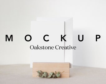 May include: A white card mockup with the text "MOCKUP Oakstone Creative" displayed on a white background. The card is propped up on a wooden stand with a sprig of greenery.