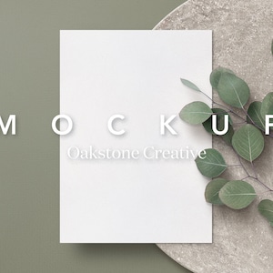 Könnte beinhalten: Ein weißer Karten-Mockup mit dem Text "MOCKUP Oakstone Creative" auf grünem Hintergrund mit einem Zweig Eukalyptusblätter auf einer Steinoberfläche.