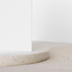 May include: A white, round, textured pedestal with a white square on top. The pedestal is made of a light-colored material, possibly concrete or stone.