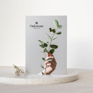 May include: A white card with the text "Oakstone Creative" and a sprig of green eucalyptus leaves held by a hand.