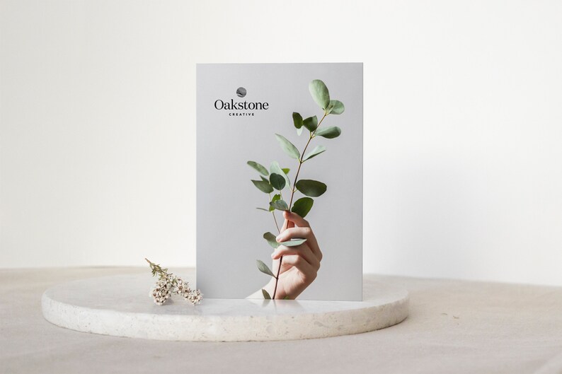 May include: A white card with the text "Oakstone Creative" and a sprig of green eucalyptus leaves held by a hand.